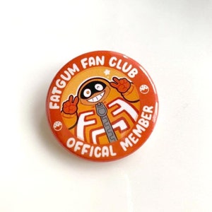 May include: A round, orange button with the text "FATGUM FAN CLUB OFFICIAL MEMBER" in white. The button features a cartoon character in an orange outfit with a zipper and peace signs.