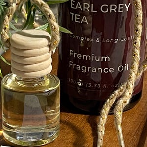 May include: A car air freshener set with a small glass bottle and a wooden cap, alongside a larger bottle labelled "EARL GREY TEA Premium Fragrance Oil". The set includes a decorative rope and a card with "Car Diffuser Instructions". The larger bottle is 100ml.