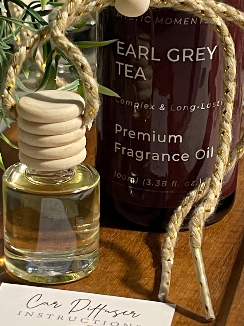 May include: A car air freshener set with a small glass bottle and a wooden cap, alongside a larger bottle labelled "EARL GREY TEA Premium Fragrance Oil". The set includes a decorative rope and a card with "Car Diffuser Instructions". The larger bottle is 100ml.