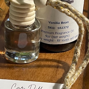 May include: A car air freshener with a wooden top and a clear glass bottle. The label reads "Vanilla Bean, Premium Fragrance Oil, 4oz (net weight)". A braided cord with gold accents is attached. The image also shows instructions for the car diffuser.