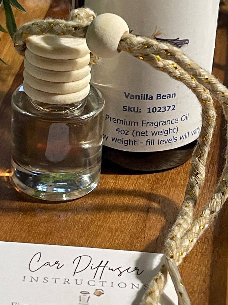 May include: A car air freshener with a wooden top and a clear glass bottle. The label reads "Vanilla Bean, Premium Fragrance Oil, 118ml (net weight)". A braided cord with gold accents is attached. The image also shows instructions for the car diffuser.
