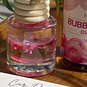 May include: A clear glass car air freshener bottle with a wooden cap and a rope. The bottle contains pink liquid and pink flowers. A second bottle with the text "BUBB" is visible. A card with the text "Car Diffuser Instructions" is in the foreground.