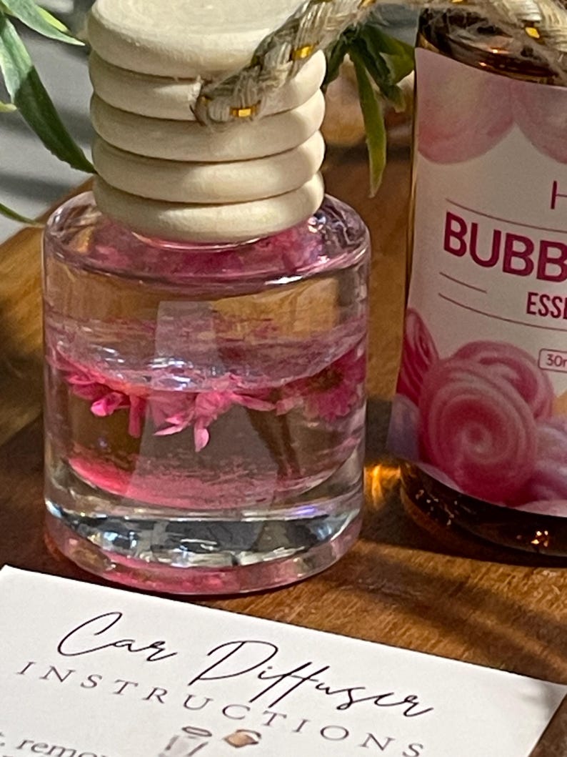 May include: A clear glass car air freshener bottle with a wooden cap and a rope. The bottle contains pink liquid and pink flowers. A second bottle with the text "BUBB" is visible. A card with the text "Car Diffuser Instructions" is in the foreground.