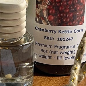 May include: A clear glass bottle with a wooden cap and a label that reads "Cranberry Kettle Corn." The label also states "Premium Fragrance" and "4oz (net weight)." A decorative rope is in the foreground.