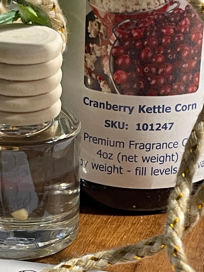 May include: A clear glass bottle with a wooden cap and a label that reads "Cranberry Kettle Corn." The label also states "Premium Fragrance" and "4oz (net weight)." A decorative rope is in the foreground.