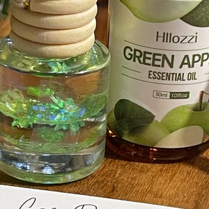 May include: A car air freshener with a wooden top and a clear glass bottle filled with a green liquid and decorative glitter. Beside it is a bottle of "Green Apple Essential Oil" with a 30ml volume. A card with "Car Diffuser Instructions" is in the foreground.