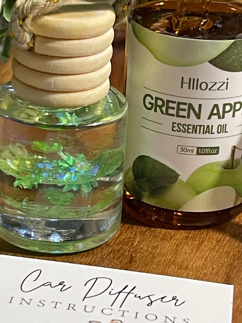 May include: A car air freshener with a wooden top and a clear glass bottle filled with a green liquid and decorative glitter. Beside it is a bottle of "Green Apple Essential Oil" with a 30ml volume. A card with "Car Diffuser Instructions" is in the foreground.