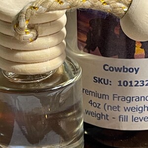 May include: Clear glass air freshener bottle with a wooden cap and a decorative rope. The label reads "Cowboy" and "4oz (net weight)". The rope is beige with gold accents.