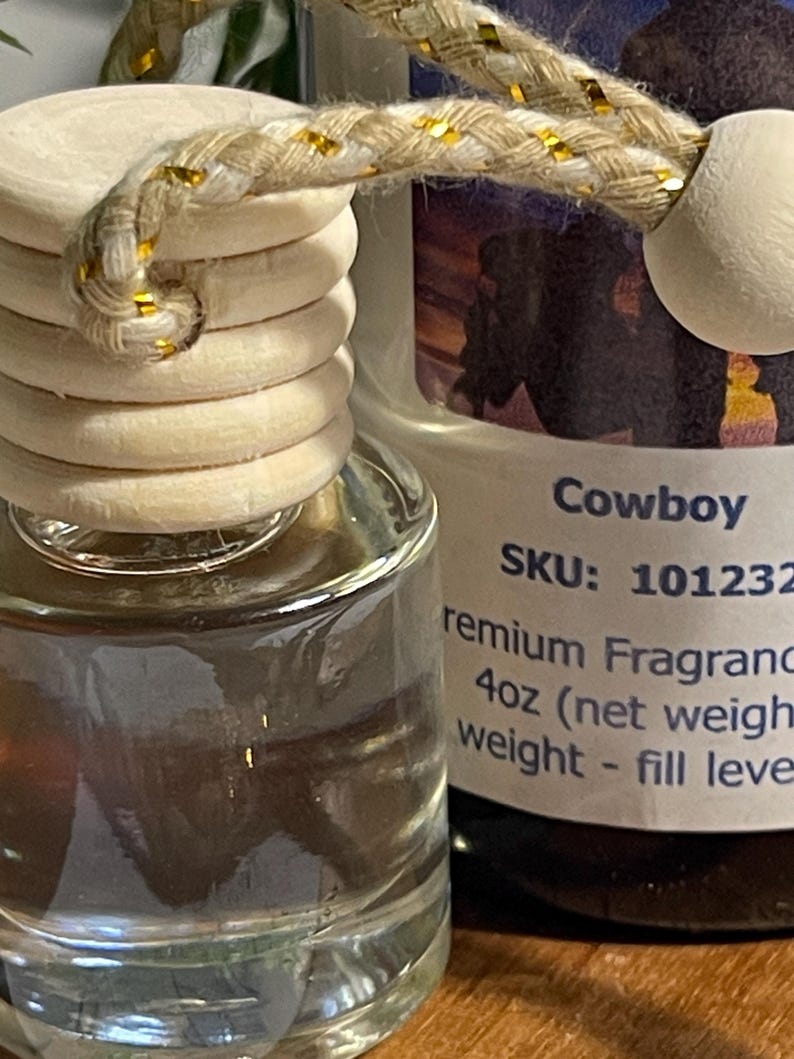 May include: Clear glass air freshener bottle with a wooden cap and a decorative rope. The label reads "Cowboy" and "4oz (net weight)". The rope is beige with gold accents.