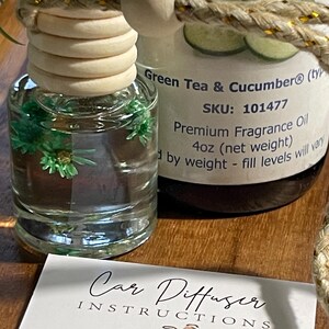 May include: A clear glass car air freshener bottle with a wooden stopper and a decorative rope. The bottle contains a green liquid with small green decorative elements. A second bottle is visible with the text "Green Tea & Cucumber". A card with instructions is in front.