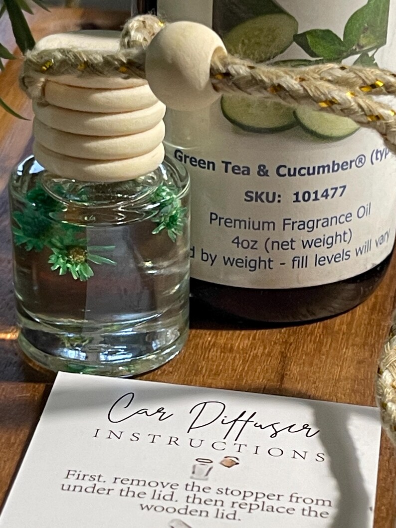 May include: A clear glass car air freshener bottle with a wooden stopper and a decorative rope. The bottle contains a green liquid with small green decorative elements. A second bottle is visible with the text "Green Tea & Cucumber". A card with instructions is in front.