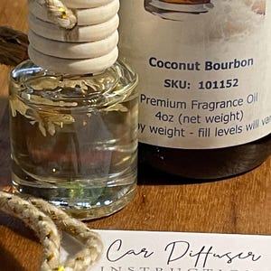 May include: A clear glass car air freshener bottle with a wooden cap and a decorative rope. The bottle contains a liquid with dried botanicals. The label reads "Coconut Bourbon" and "Premium Fragrance Oil 118ml". A card with instructions is also visible.