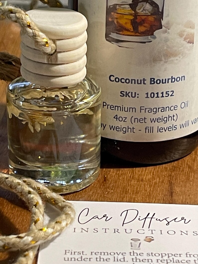 May include: A clear glass car air freshener bottle with a wooden cap and a decorative rope. The bottle contains a liquid with dried botanicals. The label reads "Coconut Bourbon" and "Premium Fragrance Oil 118ml". A card with instructions is also visible.