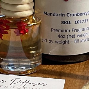May include: A glass car air freshener bottle with a wooden lid, filled with a clear liquid and red floral accents. The label reads "Mandarin Cranberry" and "Premium Fragrance". An instruction card is visible. The bottle is approximately 118 ml.