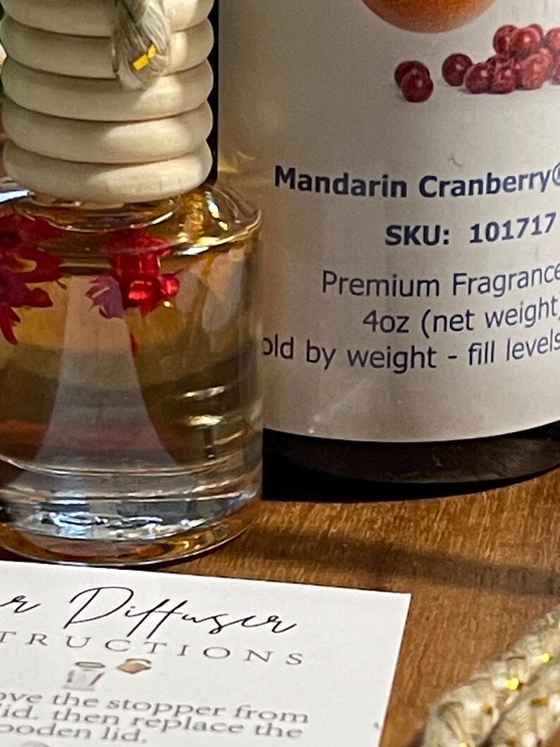 May include: A glass car air freshener bottle with a wooden lid, filled with a clear liquid and red floral accents. The label reads "Mandarin Cranberry" and "Premium Fragrance". An instruction card is visible. The bottle is approximately 118 ml.