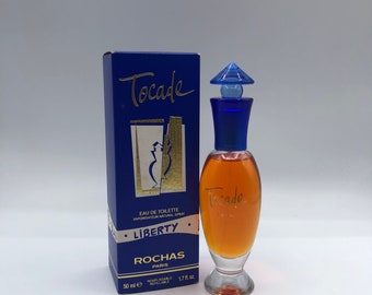 May include: A blue and gold box with the text "Tocade" and "Liberty" on it. The box contains a clear glass bottle of perfume with a blue cap and the text "Tocade" on it. The bottle is 50 ml or 1.7 fl oz.