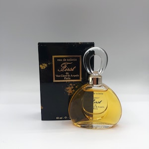 May include: A perfume bottle and box set. The bottle is a rounded shape with a clear, curved stopper. The liquid inside is amber-colored. The box is black with gold accents and the text "First de Van Cleef & Arpels Paris".