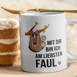 Puede incluir: Taza de cer&aacute;mica blanca con un perezoso marr&oacute;n colgado de una rama. La taza tiene texto negro que dice "Mit dir bin ich am liebsten faul" con un peque&ntilde;o coraz&oacute;n.