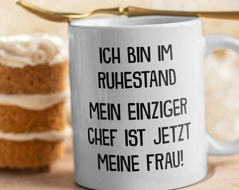 Puede incluir: Taza de café de cerámica blanca con texto negro que dice "Ich bin im Ruhestand Mein einziger Chef ist jetzt meine Frau!" La taza está sobre una superficie de madera junto a un pequeño pastel.