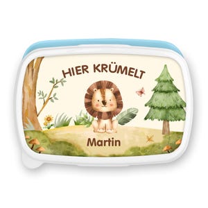 Könnte beinhalten: Eine rechteckige Brotdose mit hellblauem Deckel und weißem Rahmen. Die Vorderseite zeigt eine Aquarellillustration eines Löwenjungen in einer Waldszene mit dem Text "HIER KRÜMELT" und dem Namen "Martin".