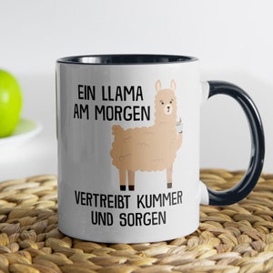 Könnte beinhalten: Weißer Keramikbecher mit schwarzem Innenraum und Henkel. Der Becher zeigt ein Cartoon-Lama, das eine Kaffeetasse hält, mit dem Text "EIN LLAMA AM MORGEN VERTREIBT KUMMER UND SORGEN" auf der Vorderseite.