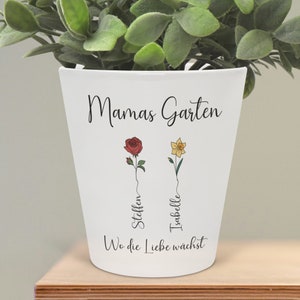 Könnte beinhalten: Weißer Keramik-Blumentopf mit dem Text "Mamas Garten" und den Worten "Wo die Liebe wächst" am Boden. Zwei Blumen sind auf den Topf gezeichnet, eine rote Rose mit dem Namen "Steffen" und eine gelbe Narzisse mit dem Namen "Isabelle".