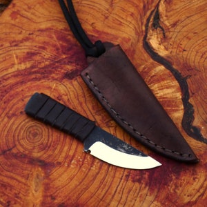 Può includere: Un piccolo coltello con manico nero e lama argentata, appoggiato su una superficie di legno. Il coltello è in un fodero di cuoio marrone con cuciture.