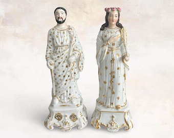 May include: Two white ceramic figurines. One figure is a man with a beard, wearing a white robe with gold star accents. The other is a woman in a white dress with a floral crown, holding a feather. Both figures stand on ornate bases.