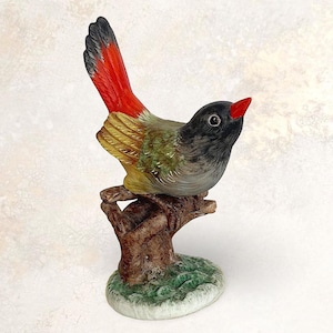 Puede incluir: Figura de pájaro de cerámica posada en una rama de árbol. El pájaro tiene cabeza negra, pico rojo y cola roja y amarilla. Las alas son verdes y amarillas. La base es un pequeño parche redondo verde y blanco.