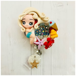May include: A white and red life preserver with a blonde cartoon girl holding onto it. The life preserver is decorated with glitter, a starfish, a crab, a seashell, and an ice cream cone. The life preserver is attached to a clear badge holder with a silver clip and a white seashell charm.