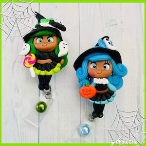 May include: Two Halloween-themed witch character badge reels. One witch has green hair, a black hat, and holds a lollipop. The other witch has blue hair, a black hat, and holds a pumpkin. Both witches are wearing black dresses and have white accents.