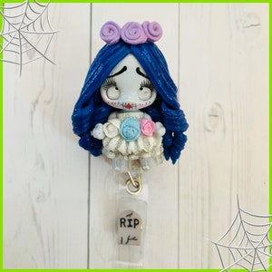 May include: A blue and white doll with a white dress and a flower crown. The doll has a stitched mouth and large eyes. The doll is attached to a clear plastic badge holder with a tombstone that says "RIP" on it.