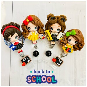 May include: Four colorful cartoon teacher figurines with brown hair and dresses, holding school supplies like a paint palette, ruler, and a chalkboard. The figurines are attached to retractable badge holders with a black bead and a small chalkboard charm with the letters "ABC" and a red apple. The text "back to SCHOOL" is written in colorful letters below the figurines.