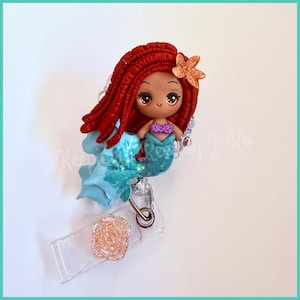 May include: A handcrafted mermaid badge reel featuring a red-haired mermaid with a purple top and teal tail. The mermaid has a glittery starfish in her hair. The badge reel has a clear plastic holder with a pink flower accent.