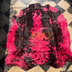 May include: A cowhide rug featuring a striking pink and black spotted design. The rug's natural form is visible, with a textured surface. The pink hue is prominent, contrasted by irregular black spots scattered across the hide.
