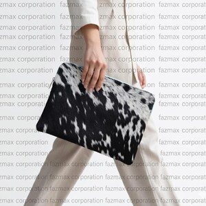 May include: A black and white cowhide clutch bag. The rectangular bag has a zip closure and is held by a person. The background has the repeated text "fazmax corporation".