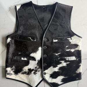 K&ouml;nnte beinhalten: Eine Weste aus schwarz-wei&szlig;em Kuhfell mit V-Ausschnitt und vier Kn&ouml;pfen. Die Weste hat zwei Vordertaschen und schwarze Ledereinfassungen. Das Kuhfellmuster ist unregelm&auml;&szlig;ig, mit schwarzen und wei&szlig;en Flecken.