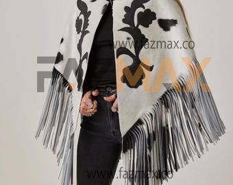 May include: A black and white cowhide poncho with black floral designs and long fringe. The poncho is worn over a black top and jeans. The website address "www.fazmax.co" is visible in the image.