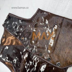 May include: A close-up of a brown and white cowhide vest with a brown leather trim. The vest has decorative silver buttons and a satin lining. The website www.fazmax.co is visible in the image.