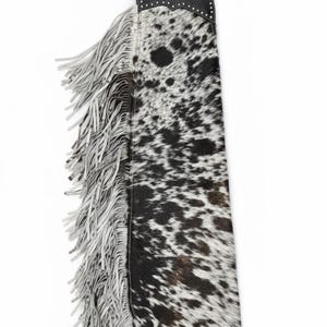 May include: Cowhide leather chaps with a black leather belt and accents. The chaps feature a spotted black and white pattern and long, white fringe along the sides. The top portion has silver-colored studs.