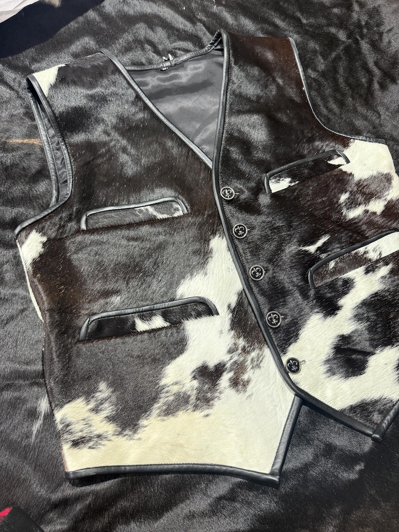 K&ouml;nnte beinhalten: Eine Weste aus Kuhfell mit einem schwarz-wei&szlig; gefleckten Muster. Die Weste hat einen V-Ausschnitt, schwarze Ledereinfassung und vier dekorative Kn&ouml;pfe. Zwei Taschen sind auf der Vorderseite sichtbar.