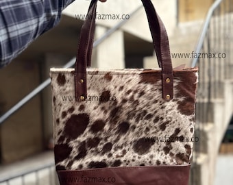 May include: A cowhide tote bag with brown leather handles and base. The bag features a spotted pattern in shades of brown and white. The handles are attached with gold-colored rivets. The website address "www.fazmax.co" is visible.