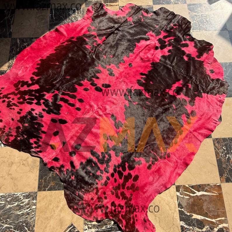 May include: A cowhide rug featuring a striking pink and black spotted design. The rug has a natural form and texture, with the website address "www.fazmax.co" printed on it. This decorative rug adds a bold statement to any room.