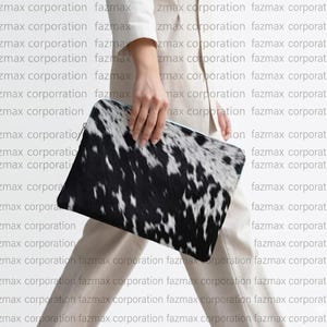 May include: A black and white cowhide clutch bag. The rectangular bag has a zip closure and is held by a person. The background has the repeated text "fazmax corporation".