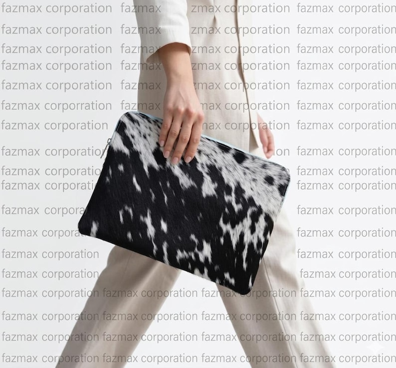 May include: A black and white cowhide clutch bag. The rectangular bag has a zip closure and is held by a person. The background has the repeated text "fazmax corporation".