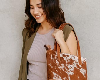 May include: A brown and white cowhide tote bag with leather handles and a leather base. The bag is being held by a person wearing a green blazer and a light purple shirt.