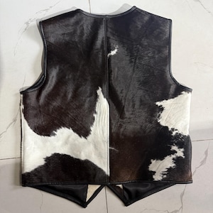 K&ouml;nnte beinhalten: Eine Weste aus Kuhfell mit schwarzem Lederbesatz. Die Weste hat ein Patchwork-Design aus schwarzem, braunem und wei&szlig;em Fell. Die Weste hat einen V-Ausschnitt und ein &auml;rmelloses Design.