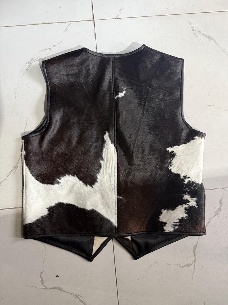 K&ouml;nnte beinhalten: Eine Weste aus Kuhfell mit schwarzem Lederbesatz. Die Weste hat ein Patchwork-Design aus schwarzem, braunem und wei&szlig;em Fell. Die Weste hat einen V-Ausschnitt und ein &auml;rmelloses Design.