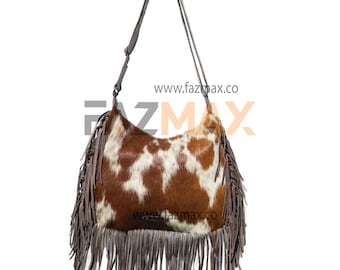 May include: A brown and white cowhide shoulder bag with a long strap. The bag has brown fringe along the bottom and sides. The text "www.fazmax.co" is printed on the bag.