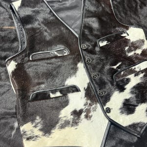 K&ouml;nnte beinhalten: Eine Weste aus Kuhfell mit einem schwarz-wei&szlig; gefleckten Muster. Die Weste hat einen V-Ausschnitt, schwarze Ledereinfassung und vier dekorative Kn&ouml;pfe. Zwei Taschen sind auf der Vorderseite sichtbar.